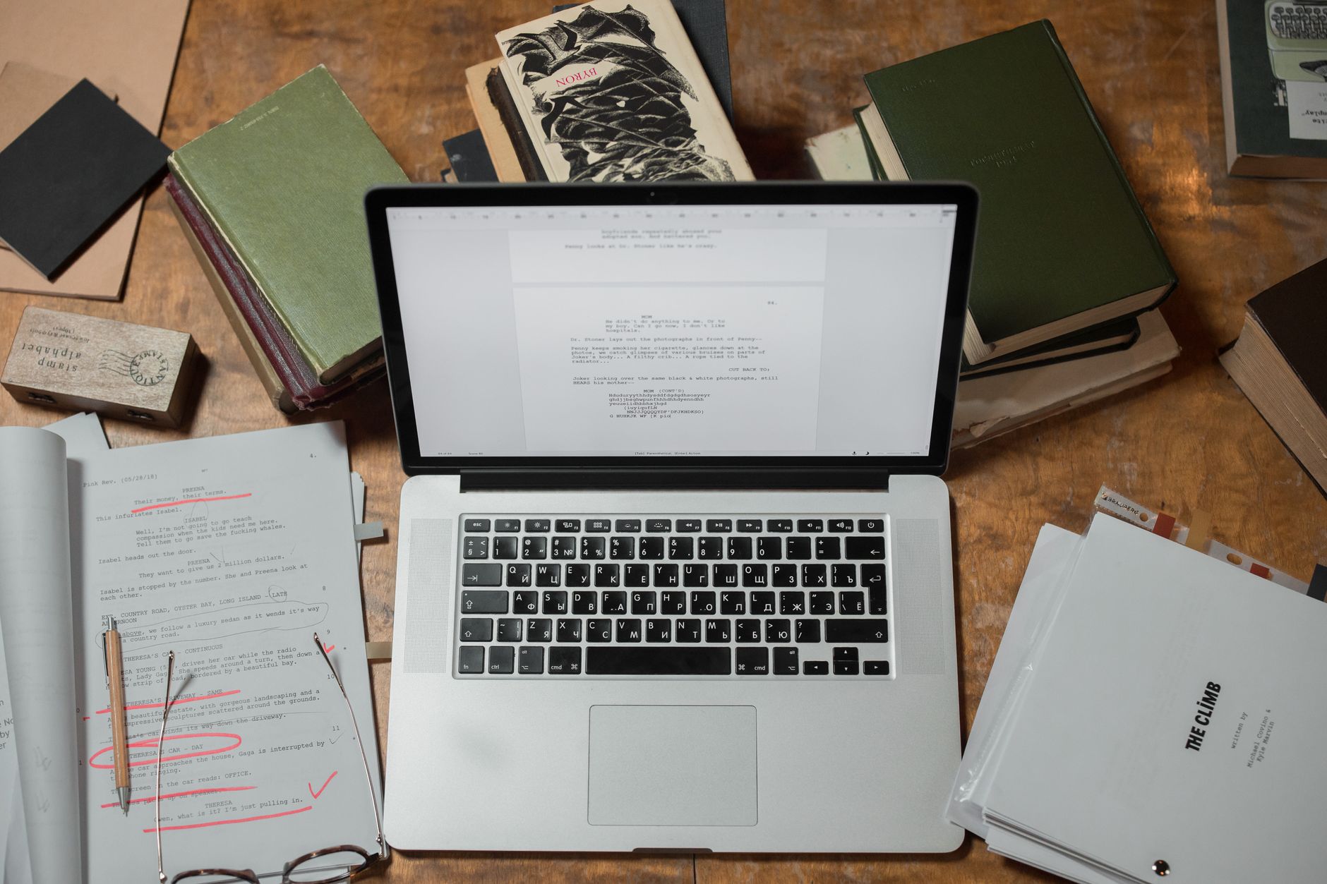 A snapshot of the founder's actual workspace from the early days. A worn oak desk with a stack of three paperback books, a legal pad with hand-drawn funnel diagrams, and an older laptop. Looks like the user snapped it themselves on a phone—imperfect angle, real environment, not a polished stock photo. No human in frame.
