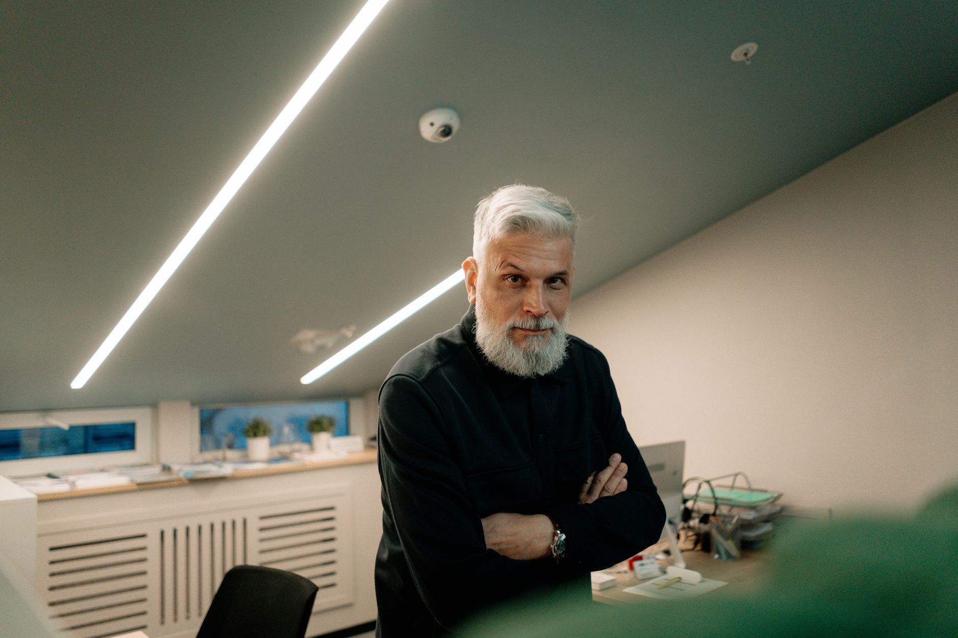 Editorial portrait of the founder, mid-40s, in a linen shirt standing in a real, slightly messy office environment (studio or garage) with morning light from a north-facing window. Shot from below at a 30-degree angle. Not in front of a rented Lamborghini. Not a fanned stack of cash.