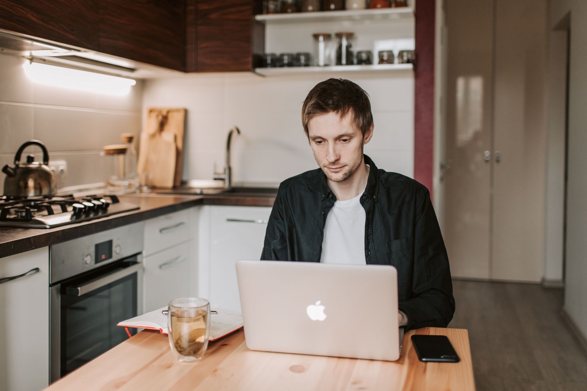 Real still of a student sitting at a kitchen table with a laptop, clean play button overlay, imperfect angle, real environment